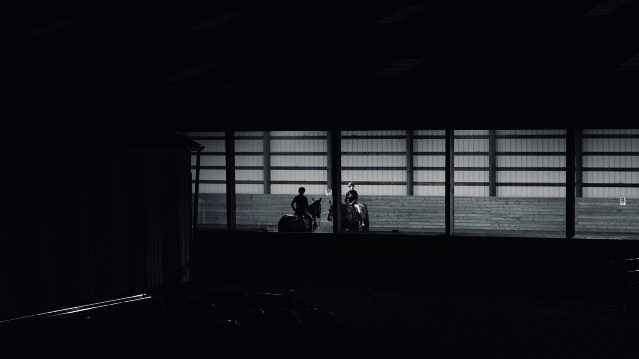 Riders silhouetted against the bright windows of a dark riding arena